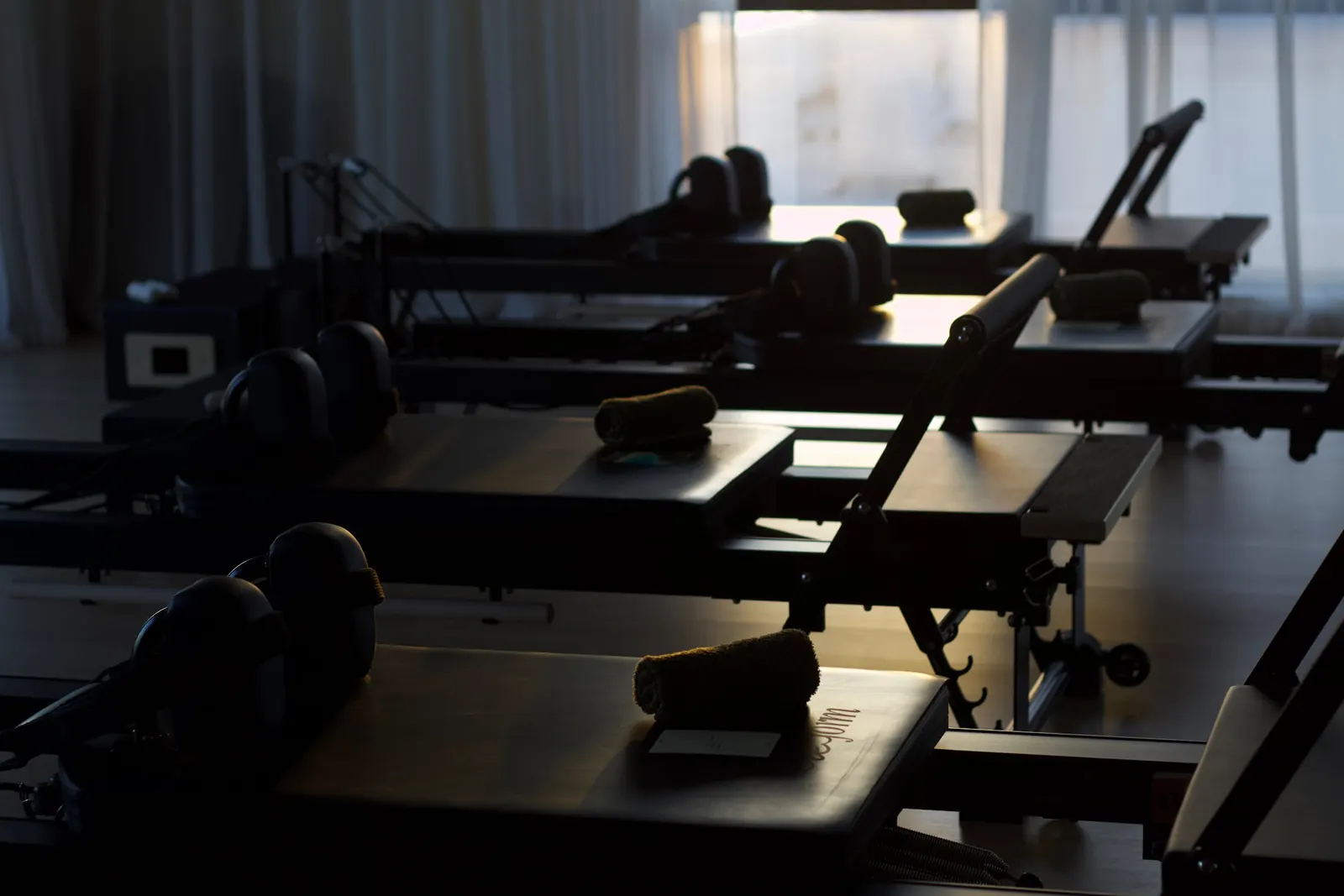 A row of reformers in the studio, backlit by soft daylight through sheer curtains